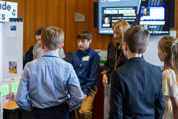 A group of fifth-grade students gather around a business project display focused on fidget spinners. The setting is an indoor exhibition space with wooden paneling and a large screen in the background showing business news.